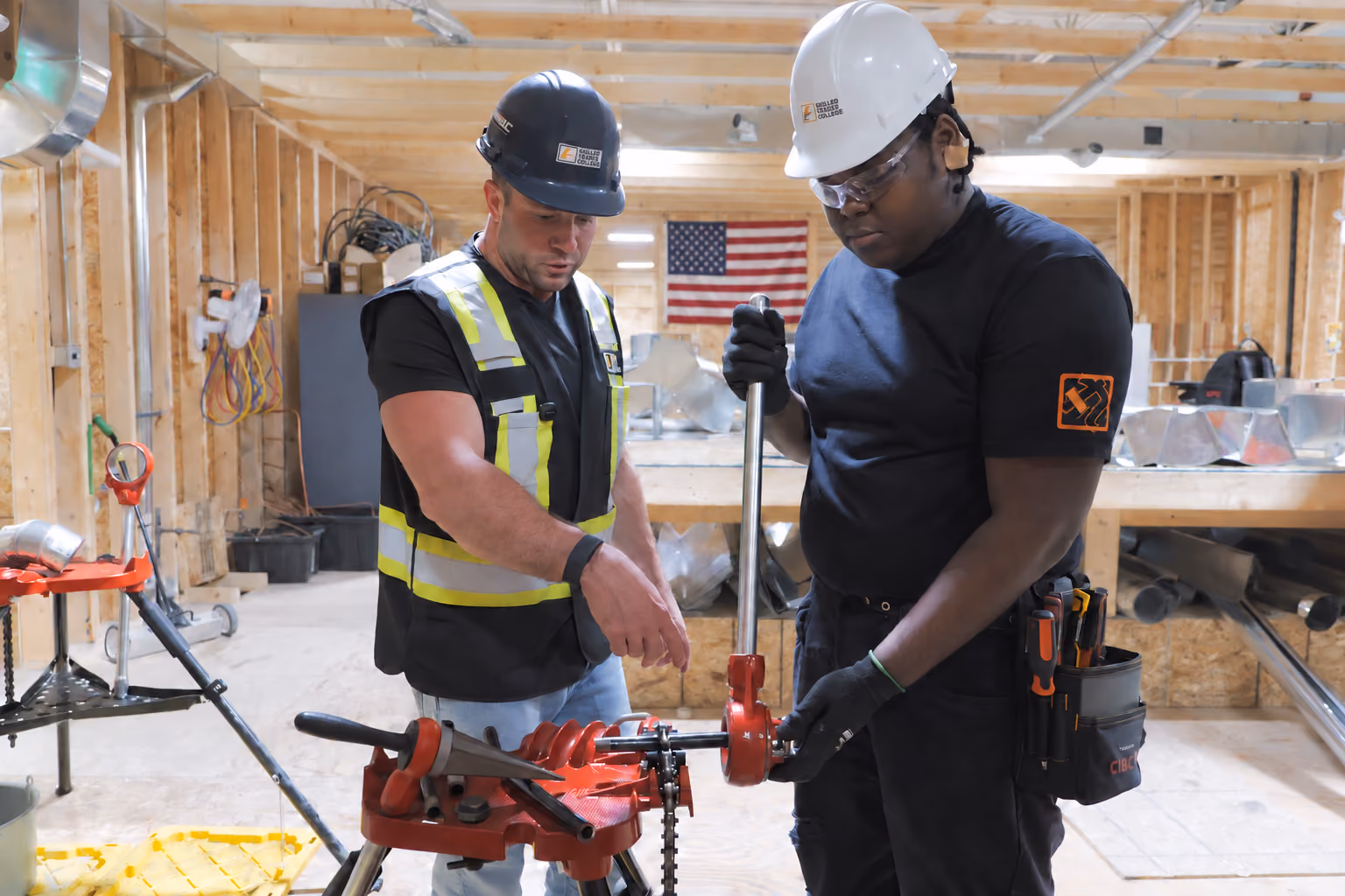Two HVAC workers wearing protective helmets and safety gear operate a red pipe threading machine inside a wooden framed workshop with an American flag on the wall in the background.