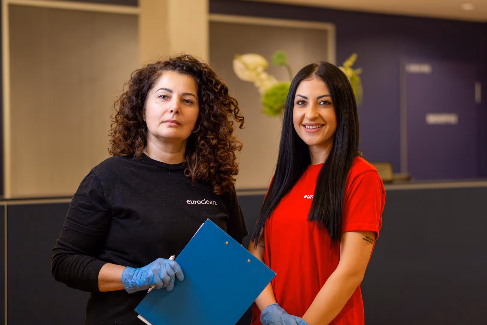 Zwei Frauen in Arbeitskleidung mit Euroclean-Logo, eine hält eine blaue Kladde, beide tragen blaue Handschuhe.