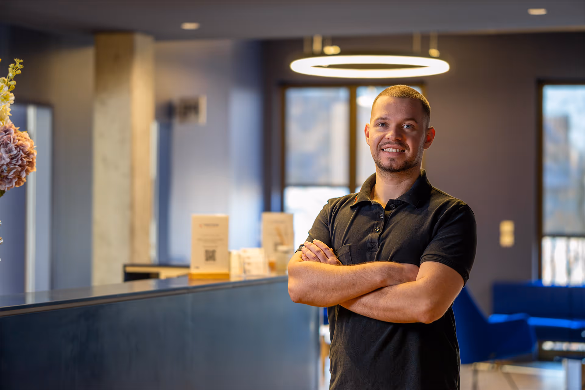 Junger Objektleiter von Euroclean mit kurzem Haar und Bart lächelnd mit verschränkten Armen in einem modernen Büro mit blauen Sesseln und großem Fenster im Hintergrund.