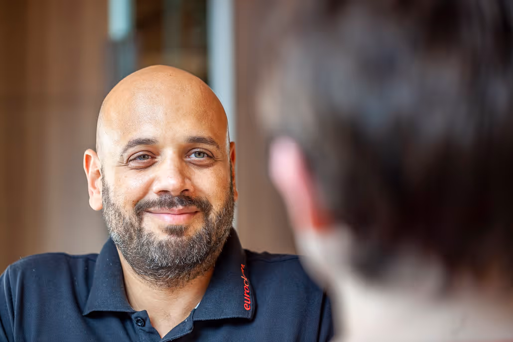 Nahaufnahme eines lächelnden Mannes mit Glatze und Bart in einem schwarzen Hemd, der eine andere Person anschaut.