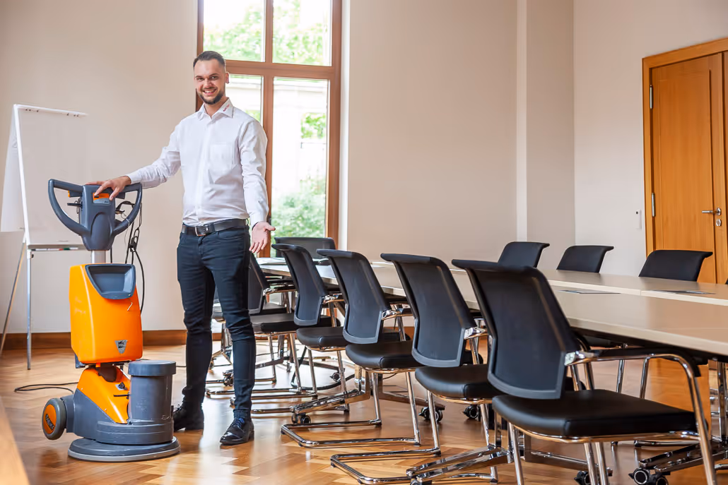 Mann in Weißhemden steht in einem Konferenzraum neben einer orangefarbenen Bodenreinigungsmaschine.