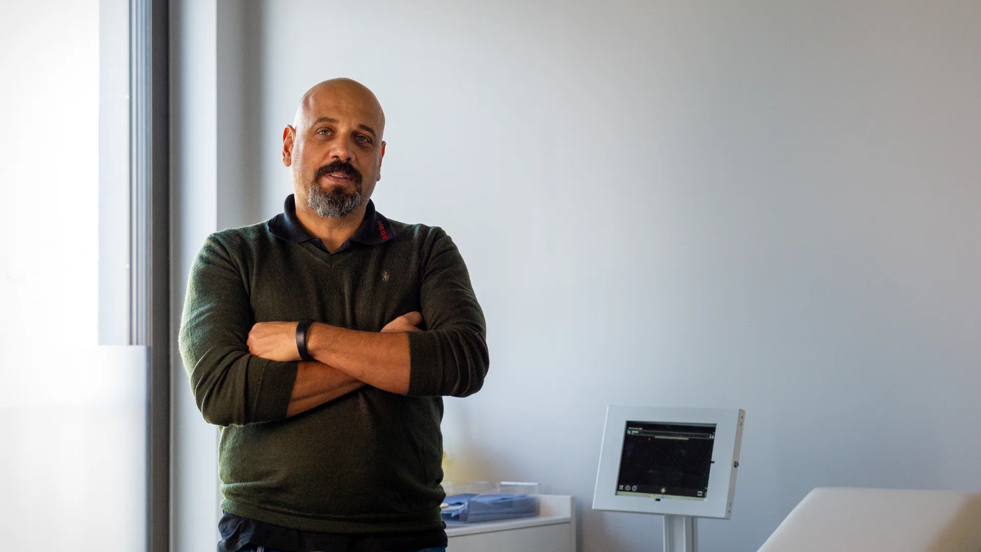 Geschäftsführer Goran Sperac mit schwarzem Pullover und blauer Jeans steht mit verschränkten Armen in einem hellen Raum neben einem medizinischen Monitor.