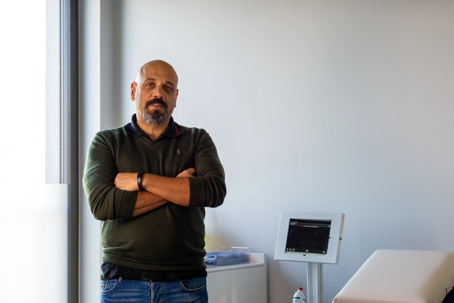 Geschäftsführer Goran Sperac mit schwarzem Pullover und blauer Jeans steht mit verschränkten Armen in einem hellen Raum neben einem medizinischen Monitor.