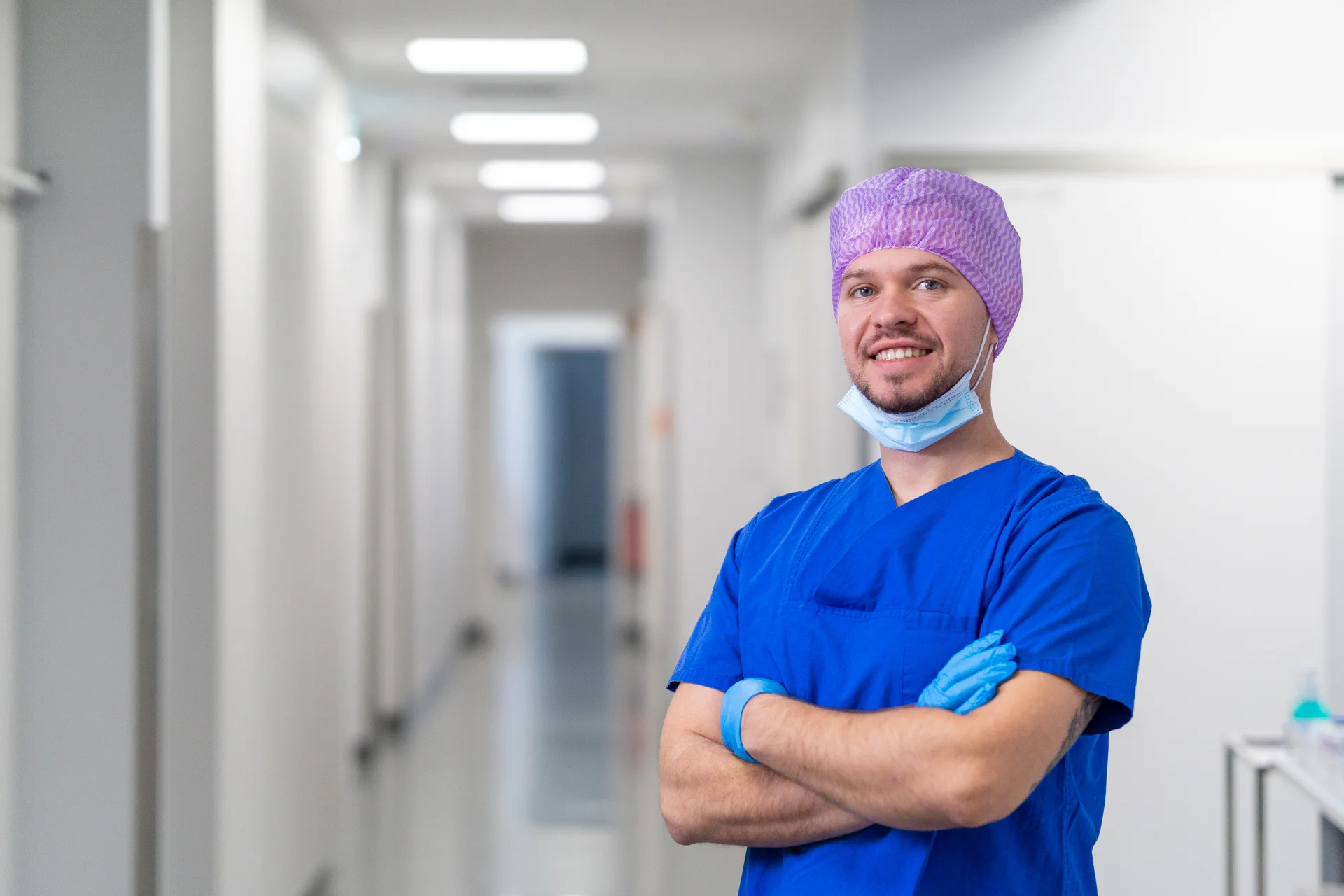 Lächelnder männlicher Objektleiter in blauer Schutzkleidung und lila Haube mit verschränkten Armen im Krankenhausflur.