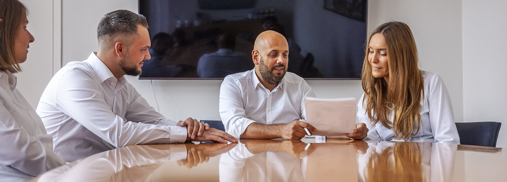 Vier Geschäftsleute in weißem Hemd, die an einem Konferenztisch sitzen, während ein Mann und eine Frau ein Dokument besprechen.