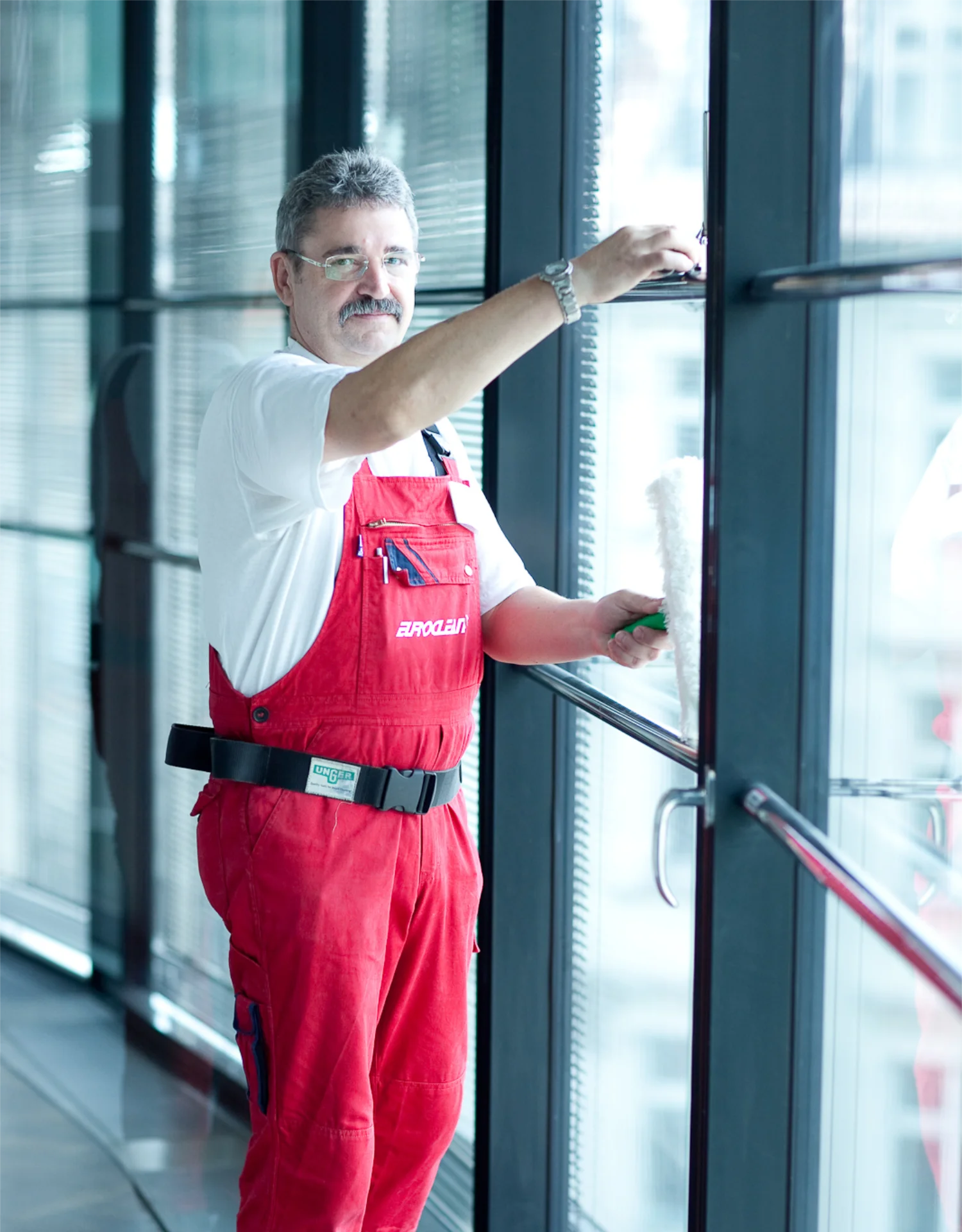 Mann in roter Arbeitskleidung reinigt Fenster mit Fensterwischer in einem modernen Büro.