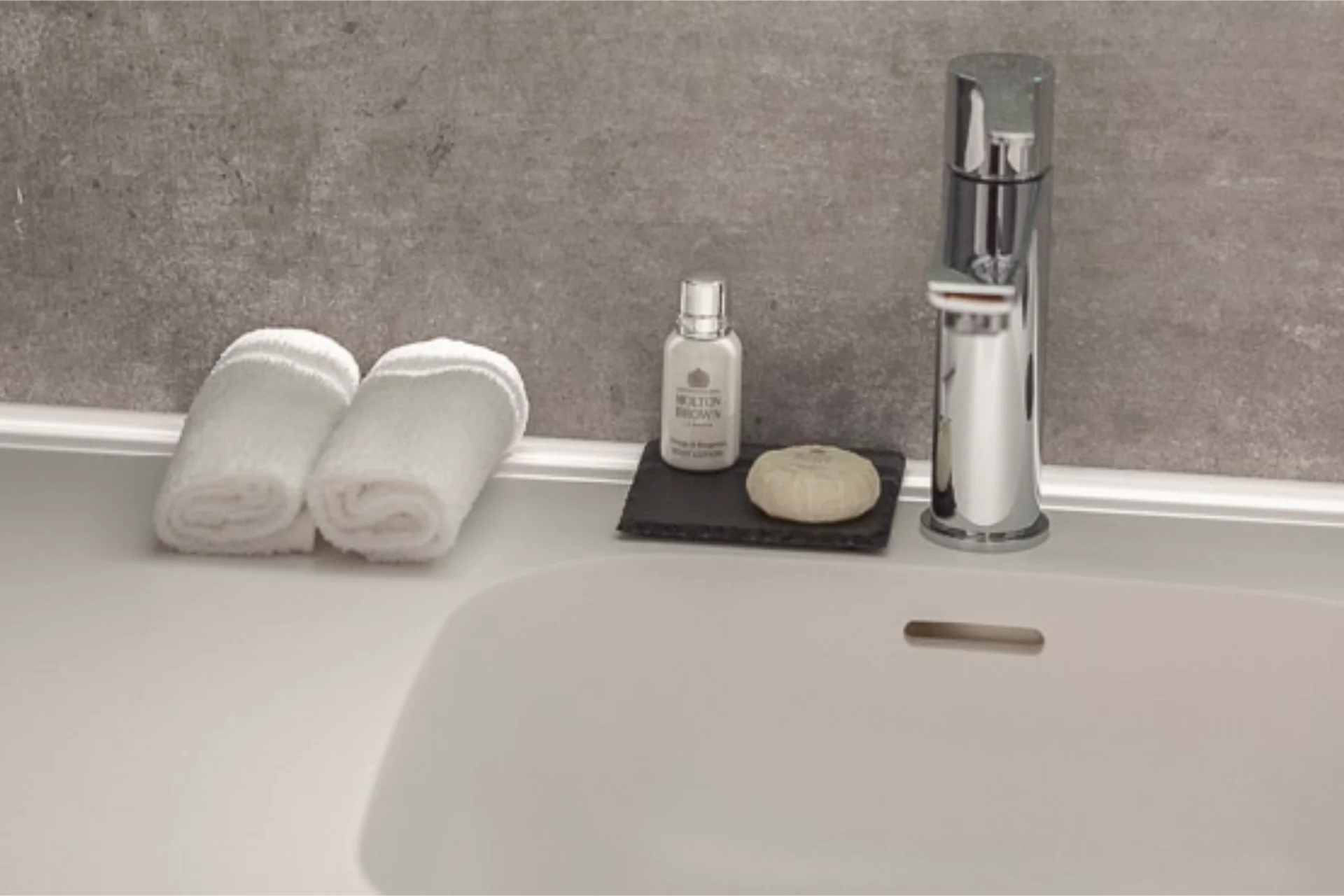 Waschbecken mit silbernem Wasserhahn, zwei zusammengerollten weißen Handtüchern, kleiner Flasche und Seifenstück auf schwarzer Ablage.