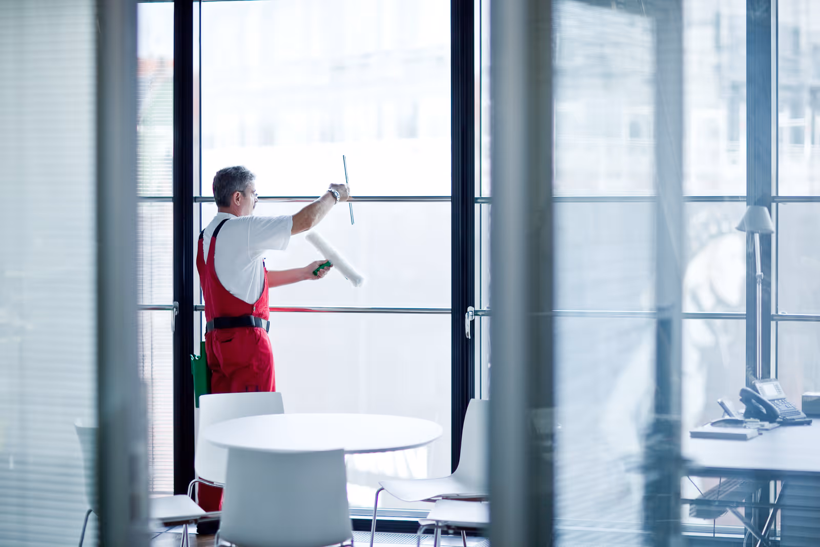 Ein Mann in roten Arbeitslatzhosen reinigt mit einem Fensterwischer und einer Rolle große Glasfenster in einem modernen Büro.
