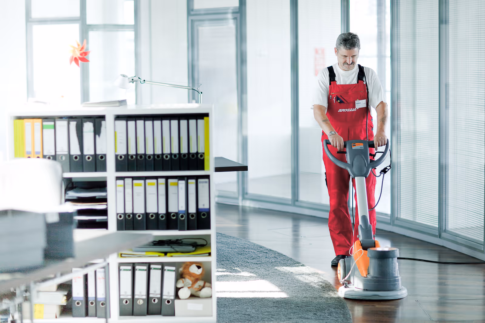 Mann in roter Arbeitskleidung reinigt Büroboden mit Poliermaschine neben Regal mit Aktenordnern und Teddybär.
