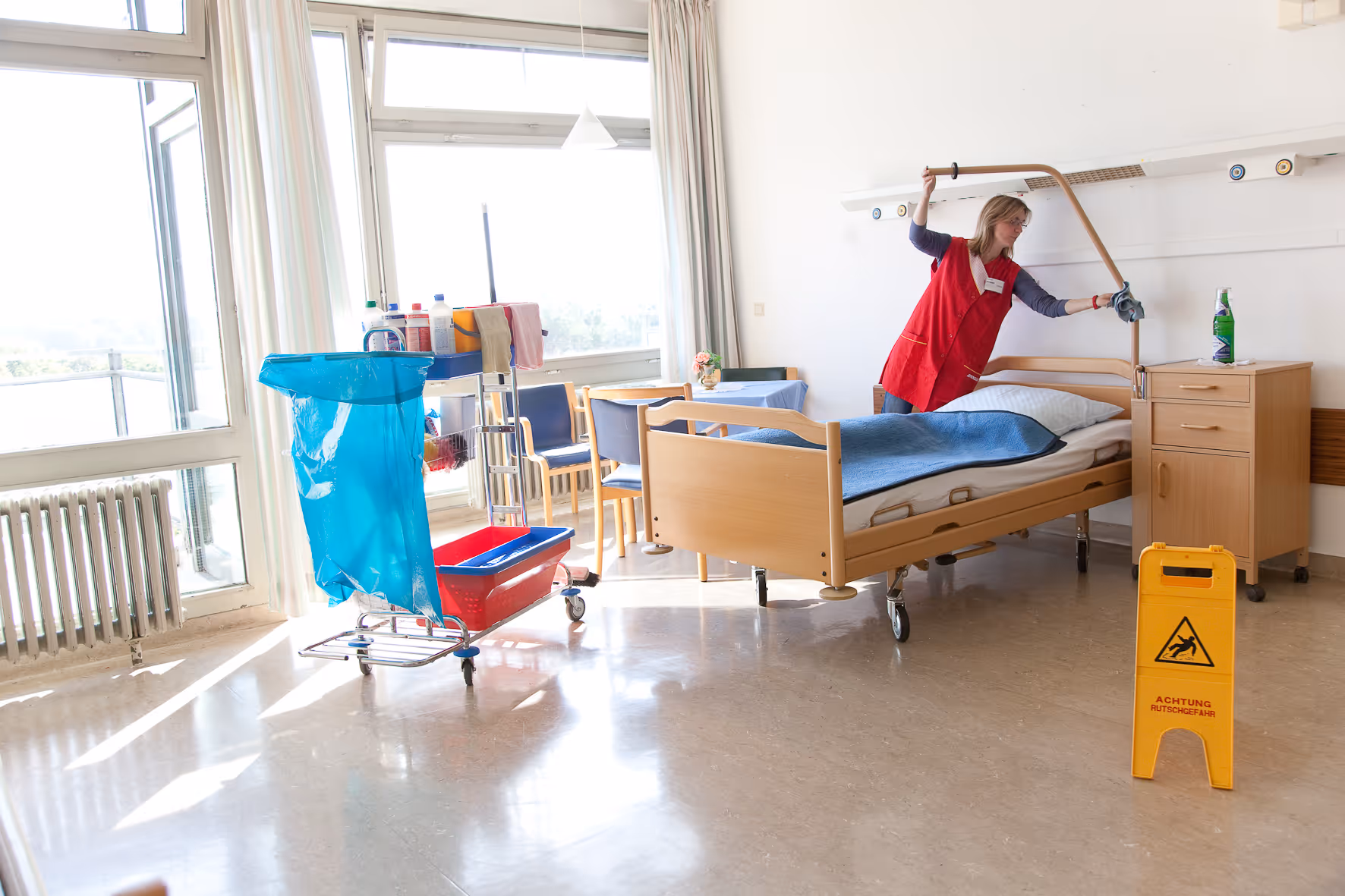 Reinigungskraft wischt in einem hellen Krankenhauszimmer das Bettgestell, neben einem Warnschild für Rutschgefahr.