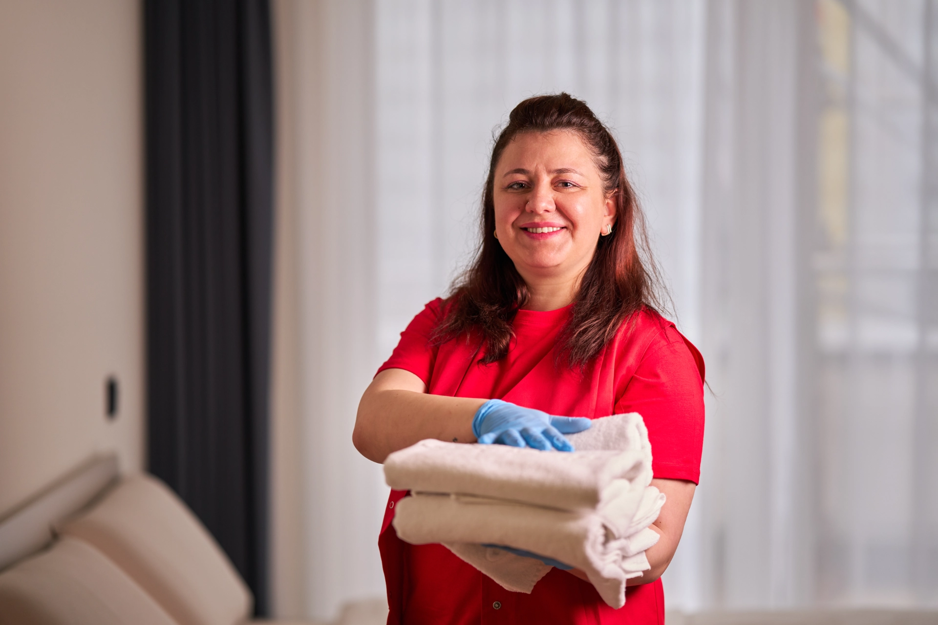 Frau in rotem Hemd und blauen Handschuhen hält gefaltete weiße Handtücher und lächelt in einem Hotelzimmer