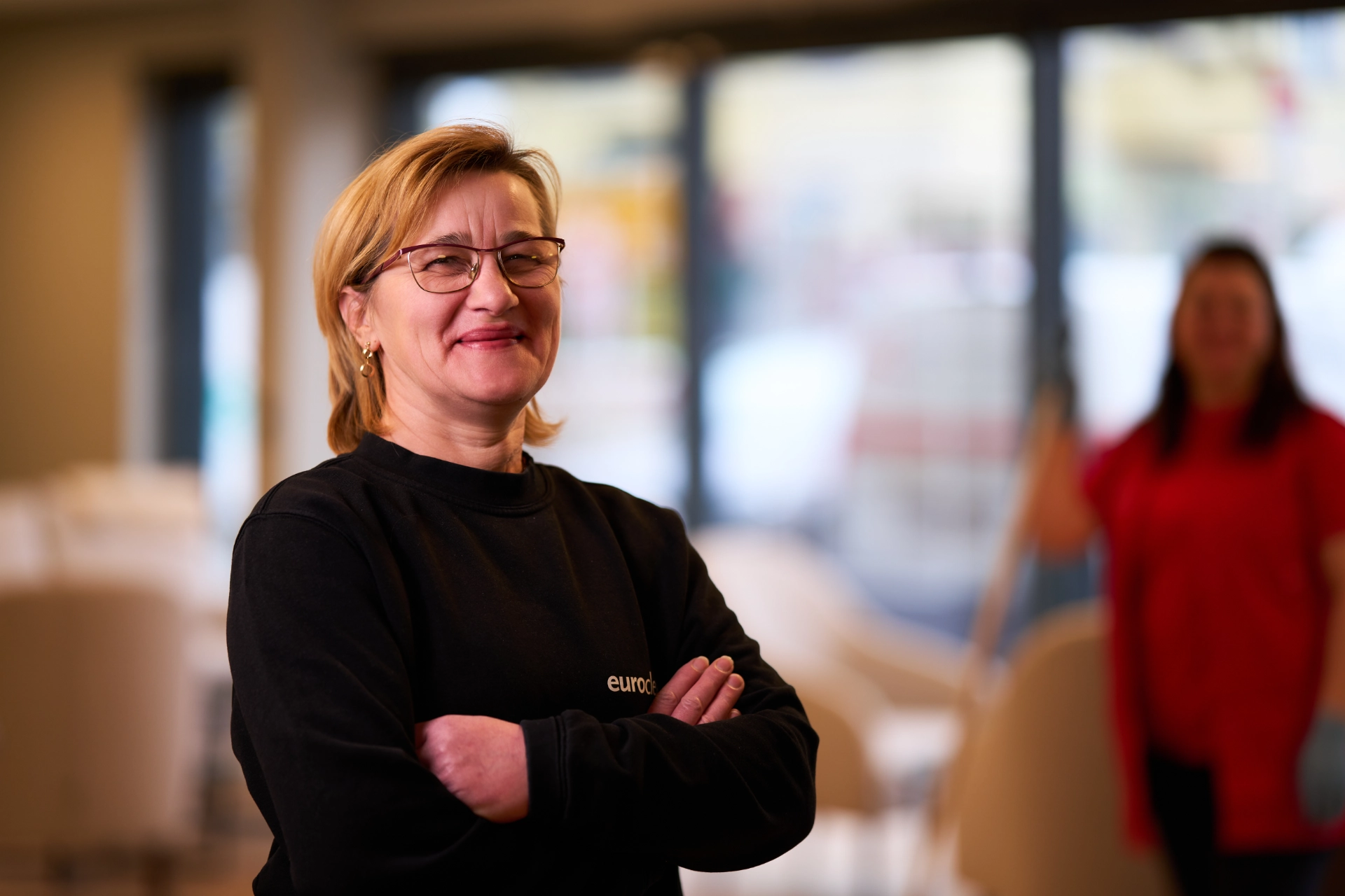 Objektleiterin mit kurzen blonden Haaren, Brille und schwarzem Sweatshirt steht lächelnd mit verschränkten Armen vor einem unscharfen Hintergrund, in dem eine weitere Person in rotem Oberteil zu sehen ist.