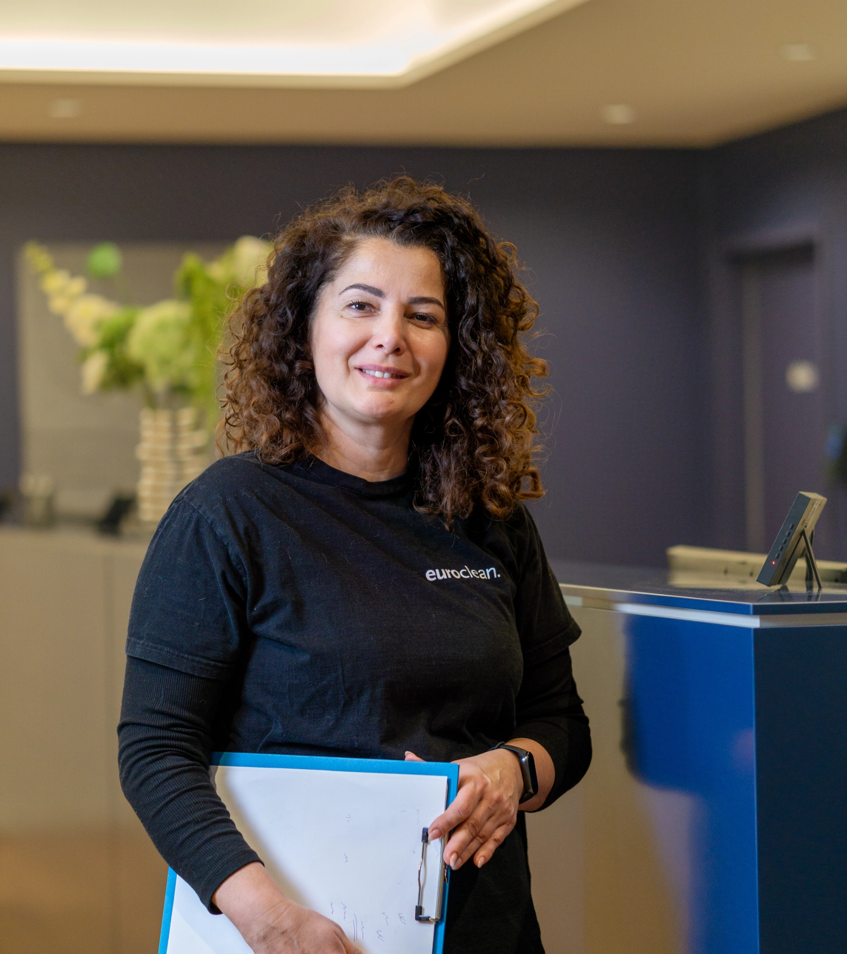 Objektleitering von Euroclean mit lockigem Haar trägt ein schwarzes Euroclean-T-Shirt und hält ein Klemmbrett in einem modernen Büro.