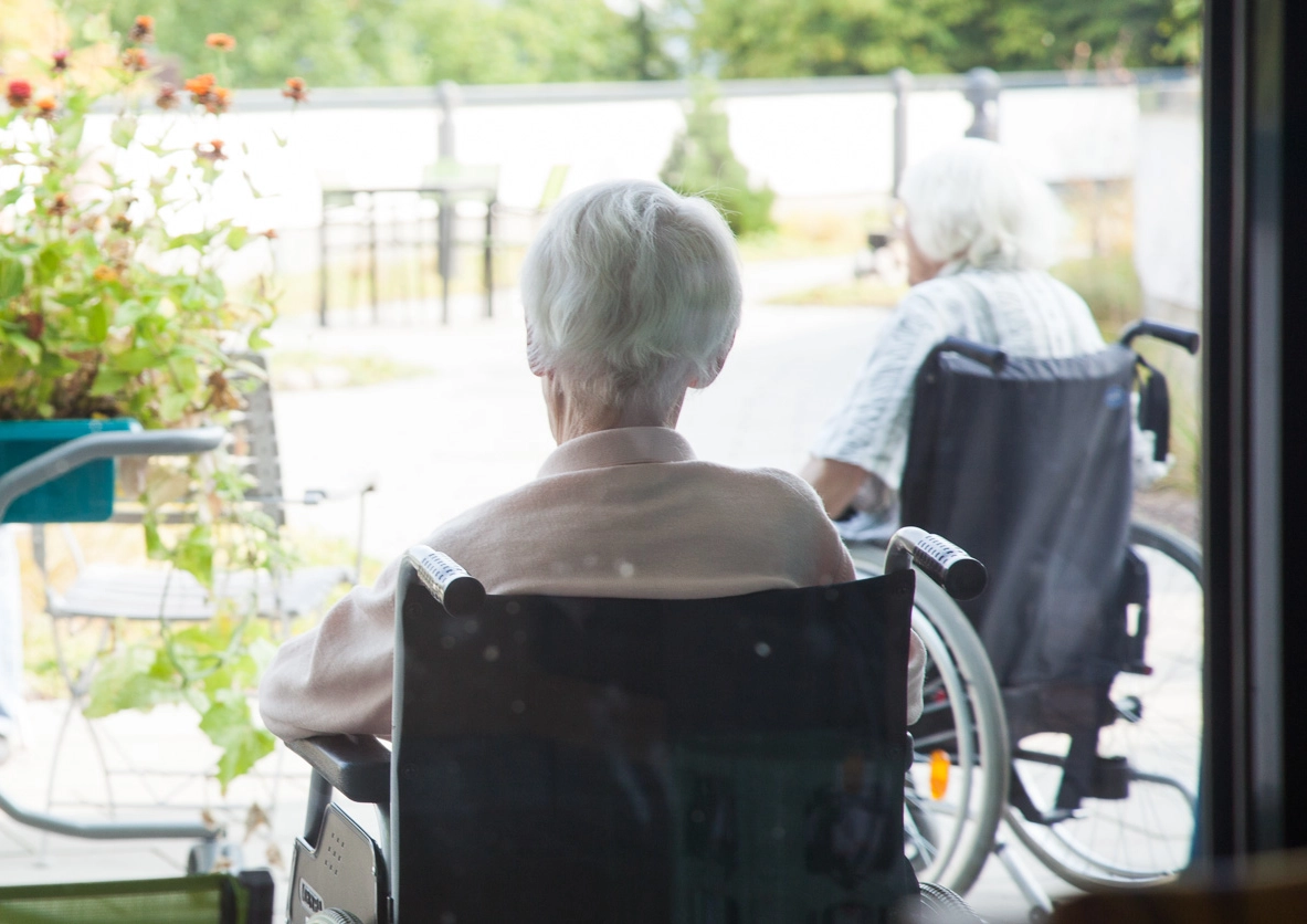 Zwei ältere Menschen mit weißem Haar sitzen im Rollstuhl auf einer Terasse neben Pflanzen und Gartenmöbeln im Altersheim.