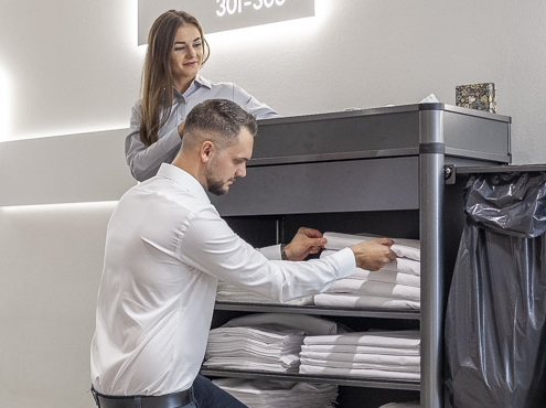 Zwei Reinigungskräfte falten weiße Handtücher auf einem schwarzen Wagen in einem hell beleuchteten Hotelgang. Der Mann hockt vor dem Wagen, die Frau im Hintergrund