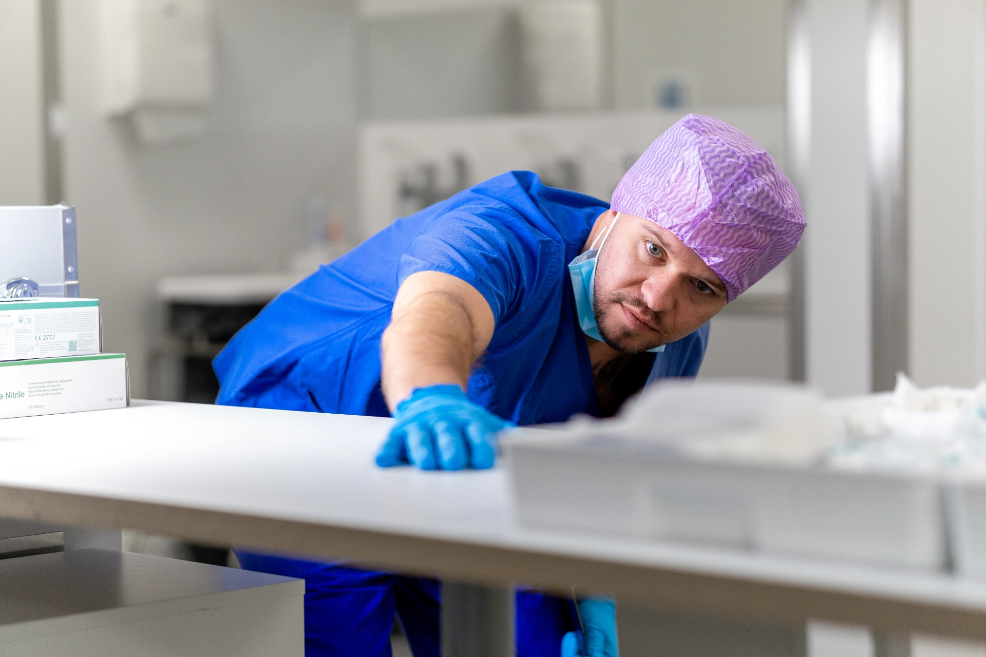 Objektleiter in blauer Schutzkleidung und lilafarbener Kappe kontrolliert konzentriert eine weiße Oberfläche in einem OP-Raum.