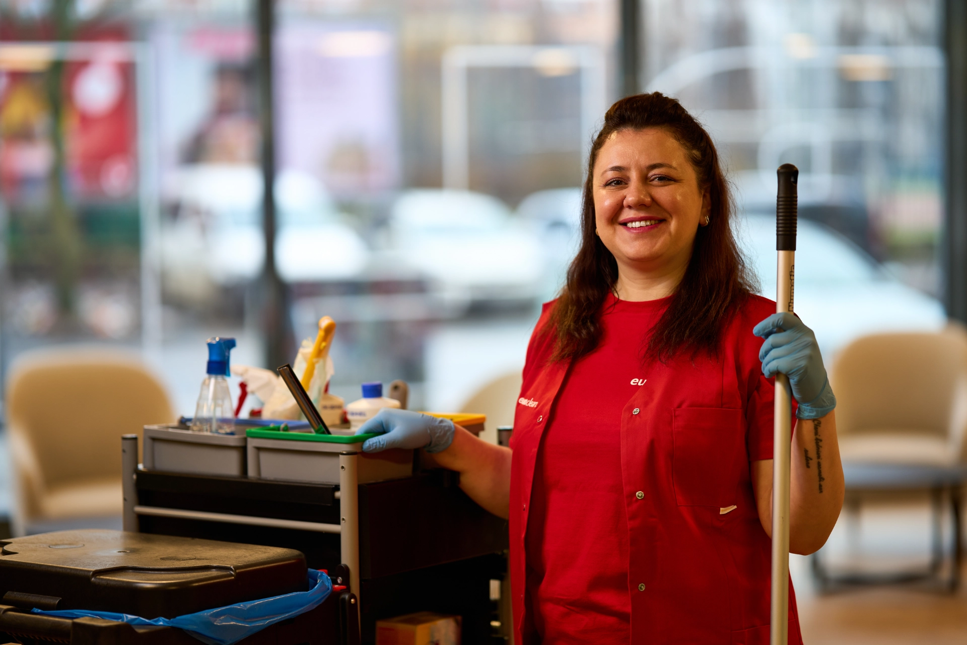 Lächelnde Reinigungskraft von Euroclean mit rotem Hemd und blauen Handschuhen steht neben einem Reinigungswagen und hält einen Mopp in einer Hotel-Lobby.