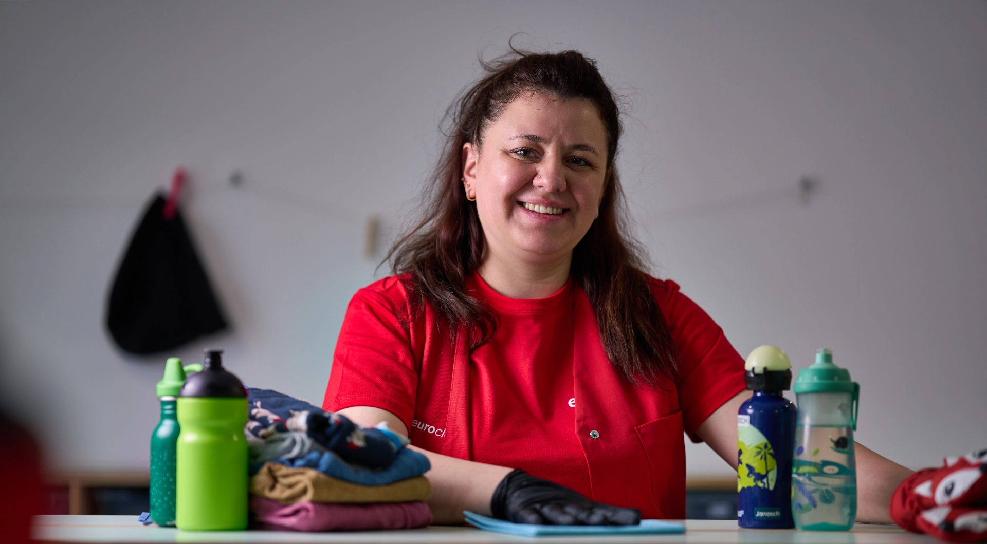 Lächelnde weibliche Reinigungskraft in rotem T-Shirt und schwarzem Handschuh wischt einen Tisch mit gefalteter Kinderkleidung und Wasserflaschen.