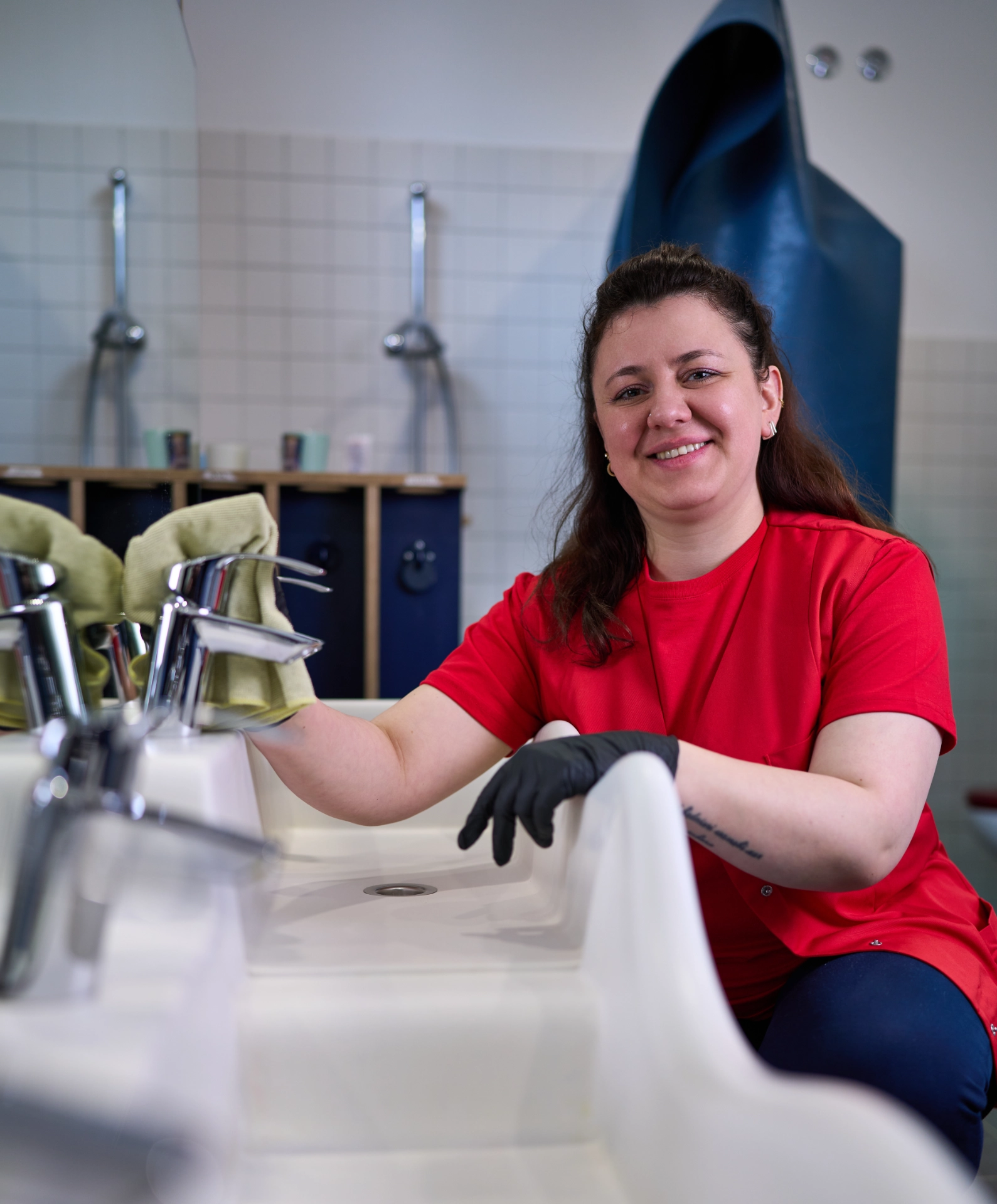 Weibliche Reinigungskraft in rotem Hemd, die ein Waschbecken mit einem grünen Tuch in einem KiTa Badezimmer wischt.