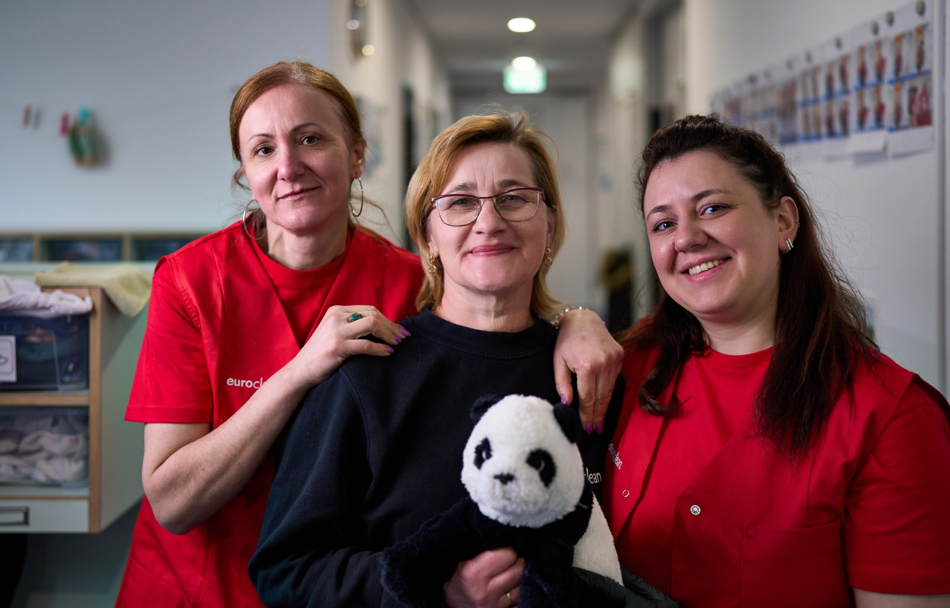Drei lächelnde Frauen von Euroclean in einem Flur, zwei tragen rote Hemden, die mittlere hält ein Plüschpanda.