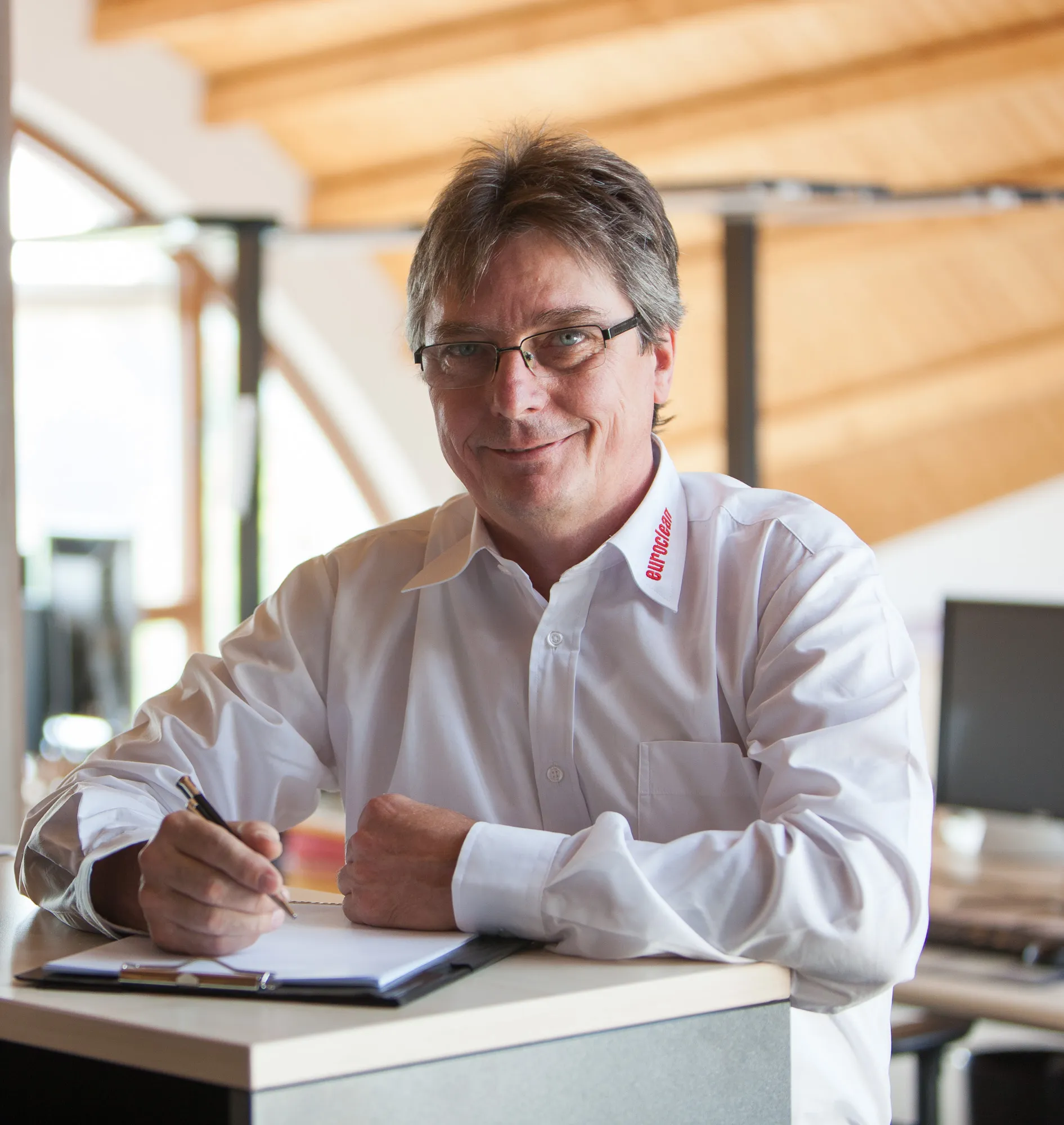 Mann mit Brille und weißem Hemd mit Schriftzug „euroclean“ auf dem Kragen sitzt an einem Schreibtisch und schreibt in ein Notizbuch.