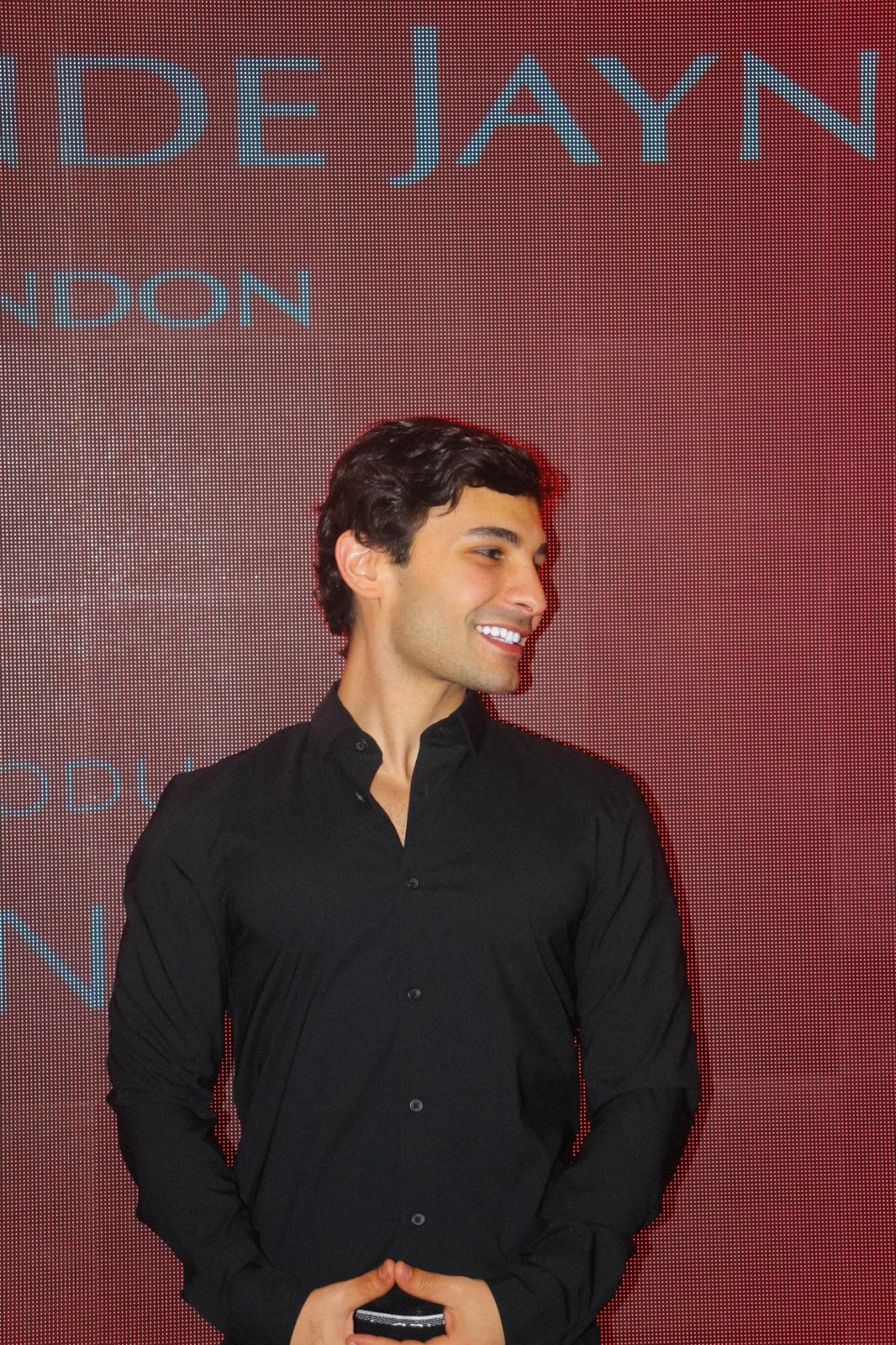 Young man with dark curly hair wearing a black button-up shirt, smiling and looking to his right against a red backdrop with partially visible light blue text.