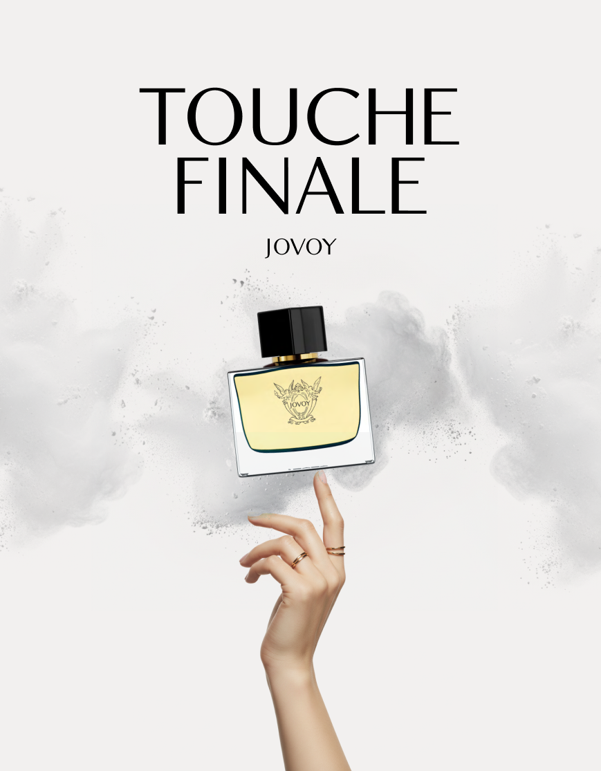 Hand with rings balancing a JOVOY Touche Finale perfume bottle against a smoky white background.