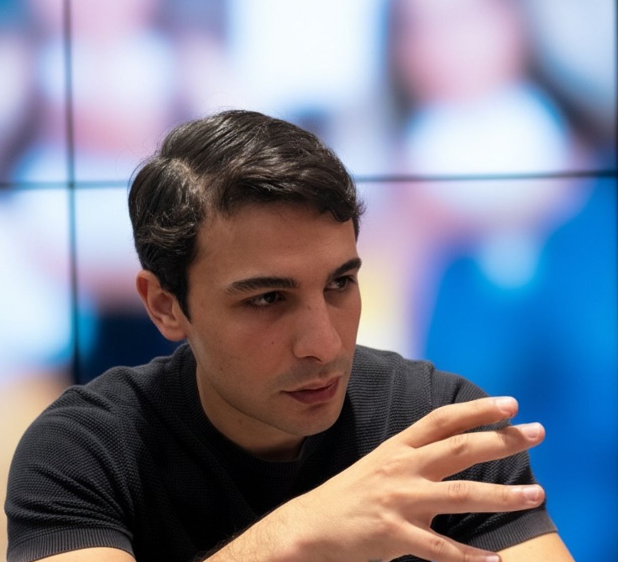 Young man in a black shirt gesturing with his hand while speaking, with a blurred colorful background.