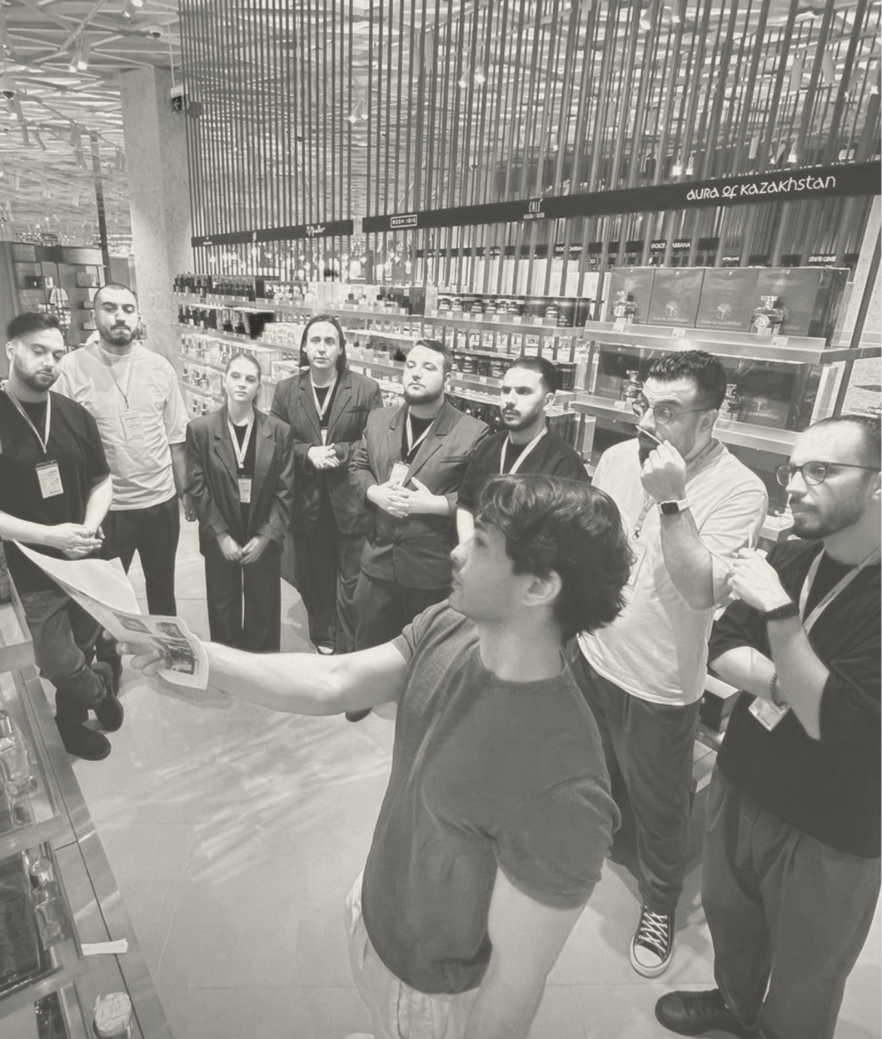 Man holding a paper explains something to a group of people standing attentively in a modern store with shelves of products.