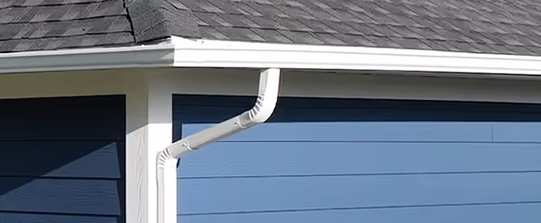 White rain gutter and downspout installed on a blue house with gray shingled roof.