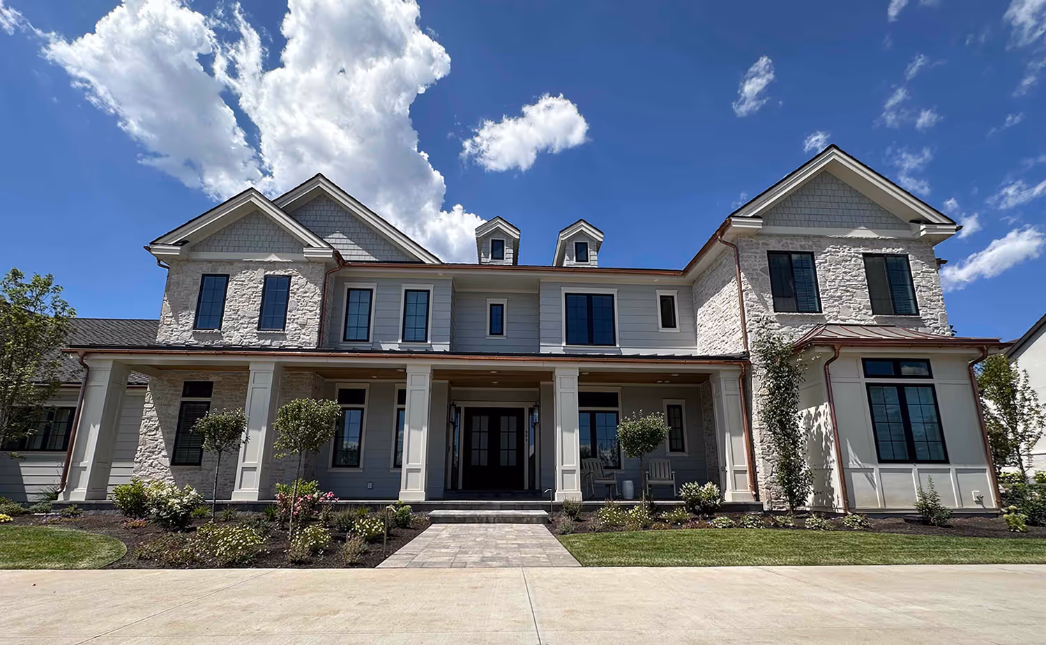 Large two-story house with stone and siding exterior, tall windows, a covered front porch, and a paved driveway under a bright blue sky with clouds.