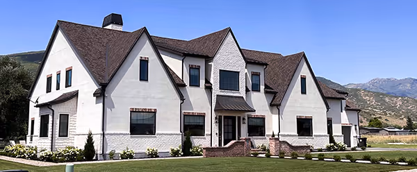 Large modern white stucco house with dark brown roof and multiple gables under a clear blue sky.
