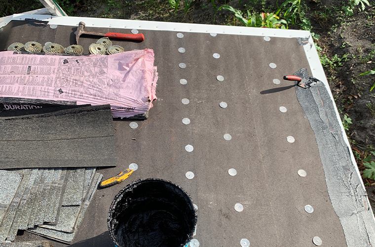 Rooftop under construction with roofing felt, tar bucket, roofing nails, and tools visible.