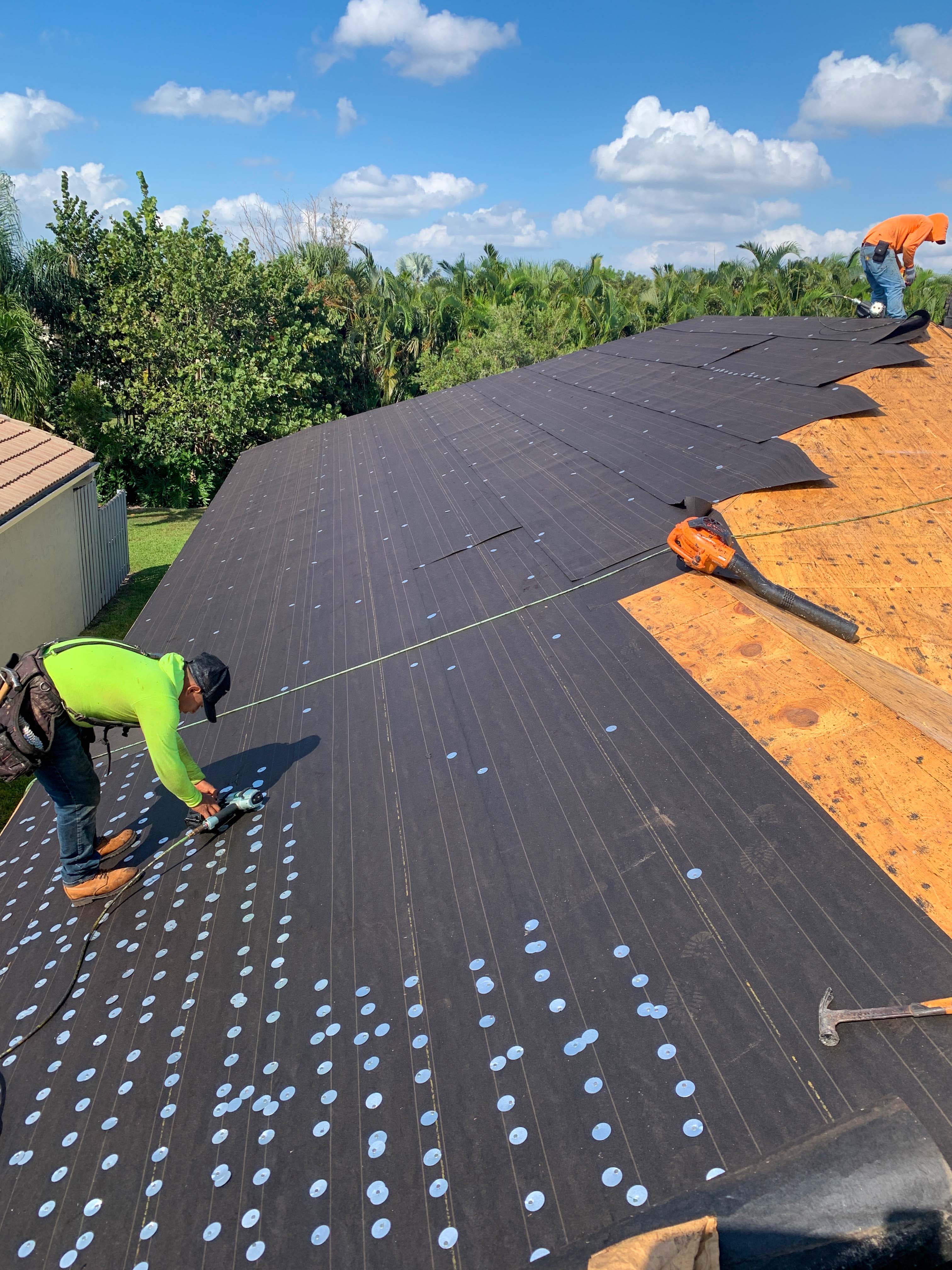 Linden Roofing workers installing roofing underlayment on a large roof under a blue sky with scattered clouds.