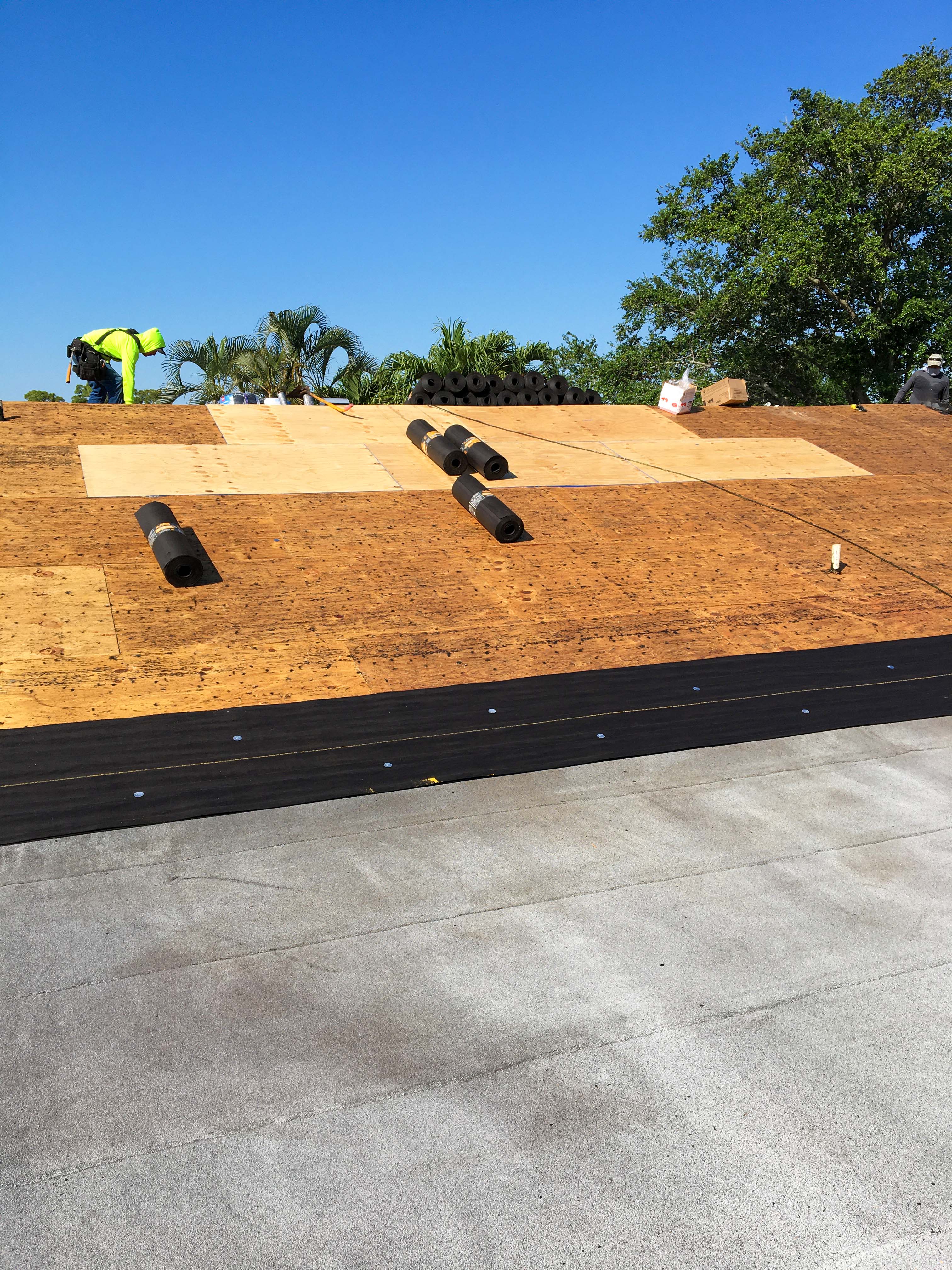 Workers installing plywood sheathing and black roofing underlayment on a roof under a clear blue sky.