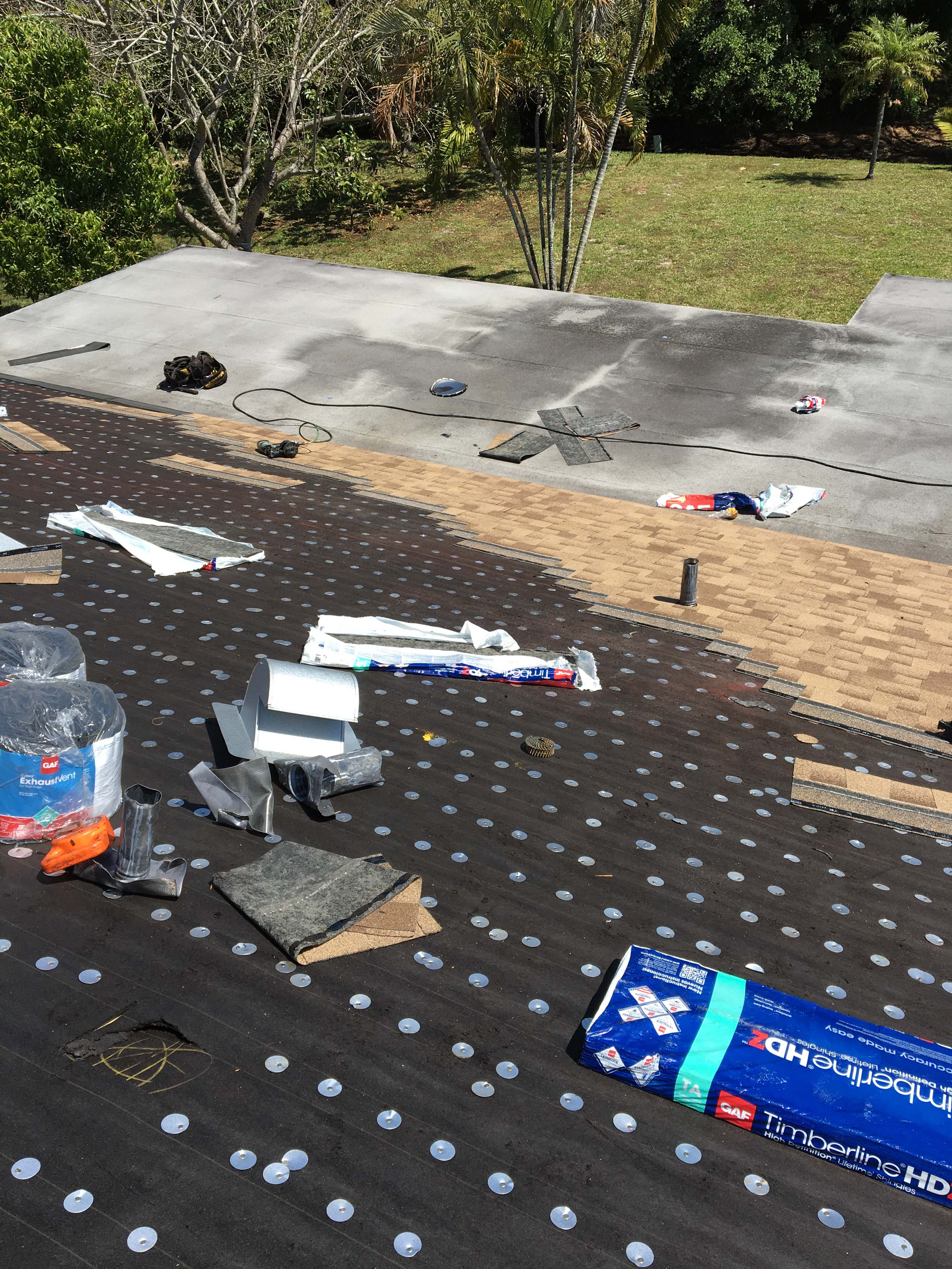 Roof under construction with black roofing underlayment secured by plastic caps and partially installed tan asphalt shingles, surrounded by roofing materials and tools.