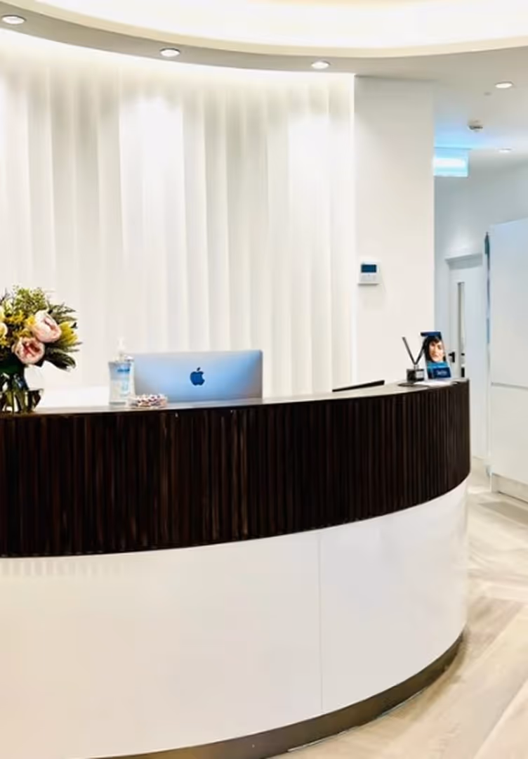 Modern curved reception desk with a silver Apple computer, a flower vase, hand sanitizer, and a small photo frame in a brightly lit office.