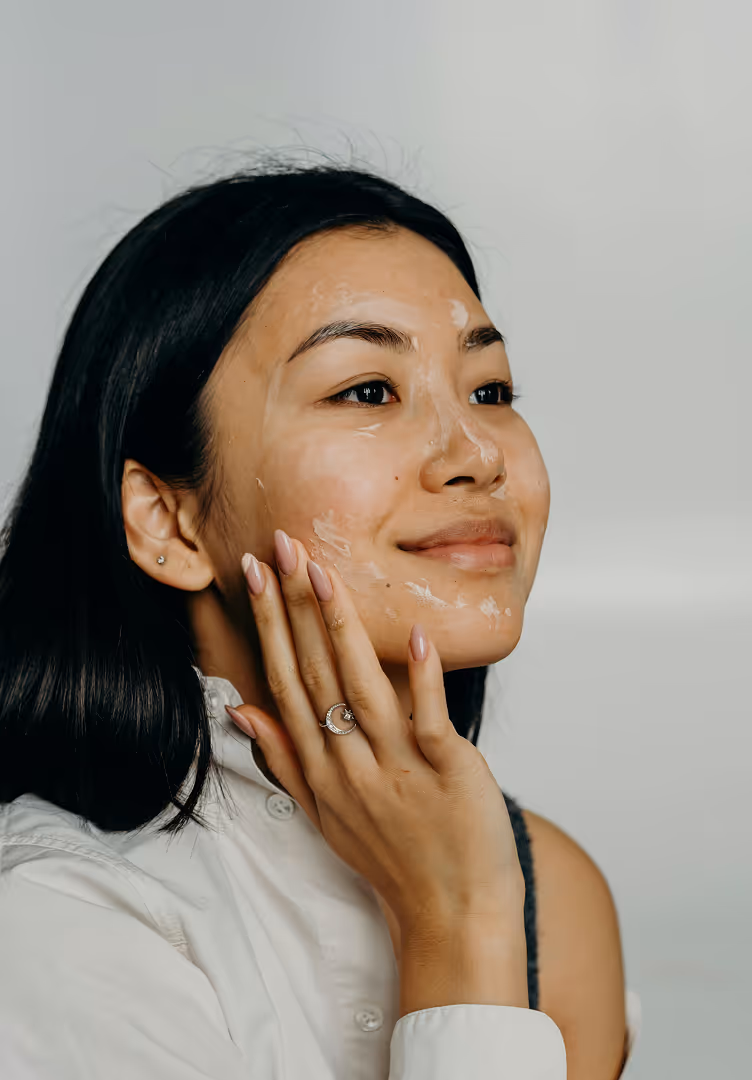 Smiling woman with black hair applying cream to her cheek, wearing a white shirt and a silver ring with a crescent moon design.