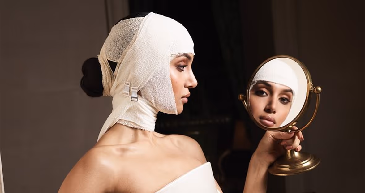 Woman with bandages wrapped around her head looking at her reflection in a handheld round mirror.