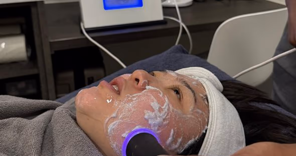 Person receiving a facial treatment with a device emitting blue light on a cream-covered cheek, lying on a towel-covered surface with a white headband.
