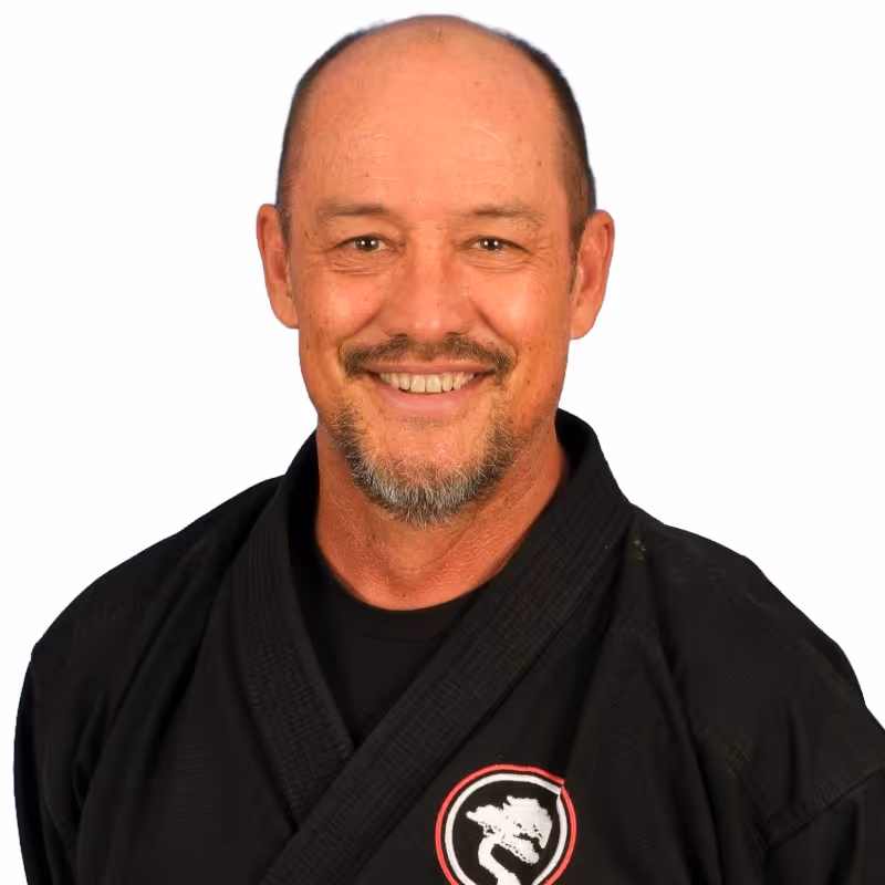 Smiling middle-aged man with a goatee wearing a black martial arts uniform in Temecula, CA at the United Studios of Self Defense Temecula dojo with a white dragon emblem on the chest.