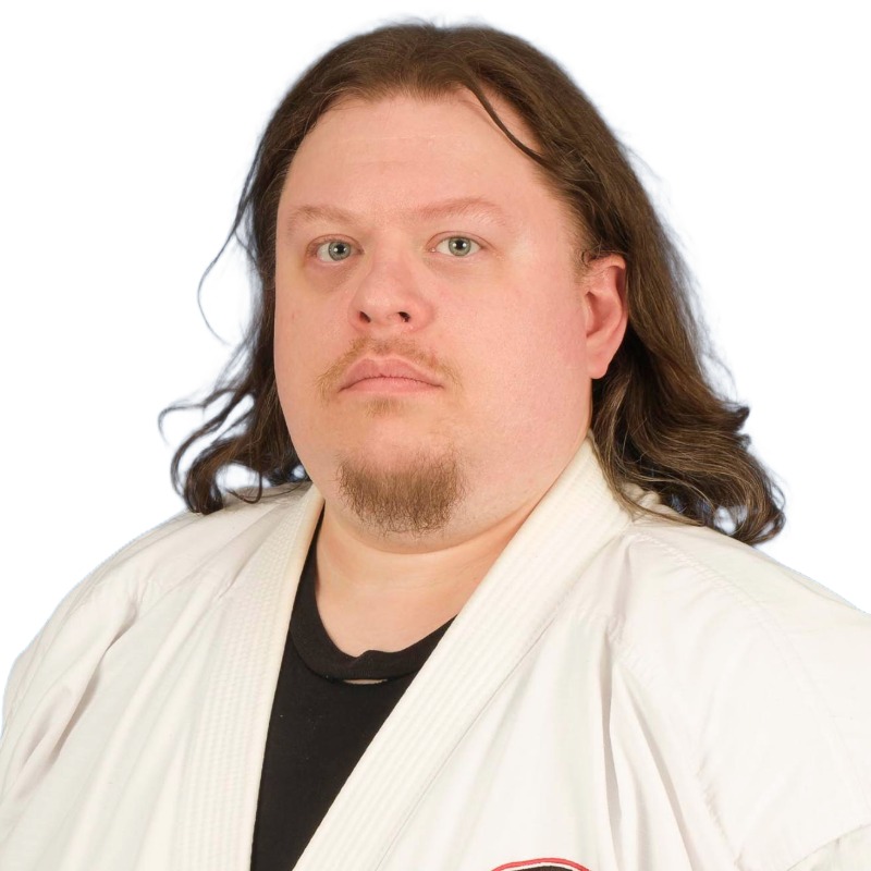 Man with shoulder-length brown hair and a goatee wearing a white martial arts gi over a black shirt in Temecula, CA at the United Studios of Self Defense Temecula dojo, looking straight ahead.
