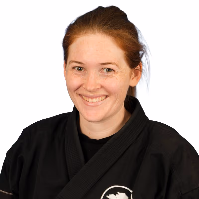 Smiling woman with red hair wearing a black martial arts uniform in Temecula, CA at the United Studios of Self Defense Temecula dojo against a white background.