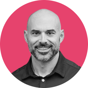 Smiling bald man with a trimmed beard wearing a dark collared shirt against a bright pink circular background.