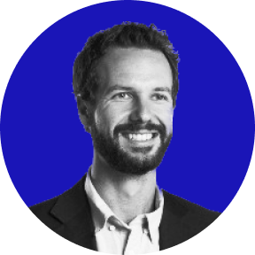 Smiling man with short curly hair and beard wearing a suit jacket and white shirt in front of a blue circular background.