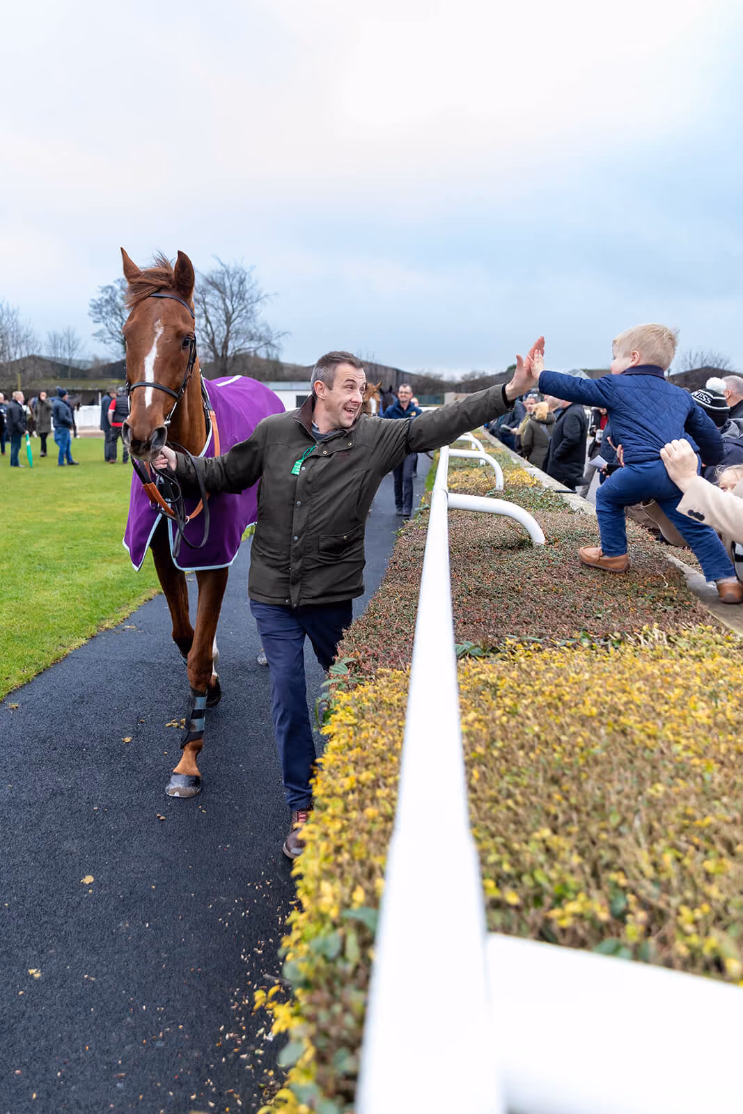 Handler leading a racehorse while high-fiving a young fan at the rail, Carlisle Racecourse — by AlTopping.com