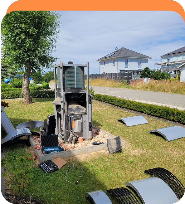 Auf dem Rasen eines Vorgartens steht ein zerlegter Außenkamin mit mehreren abgenommenen Metallverkleidungen und Werkzeugsatz daneben.