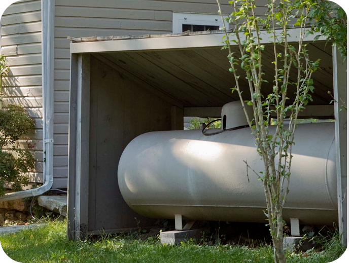 Ein großer weißer Gastank unter einem kleinen Unterstand neben einem Haus mit Garten und einem Baum im Vordergrund.