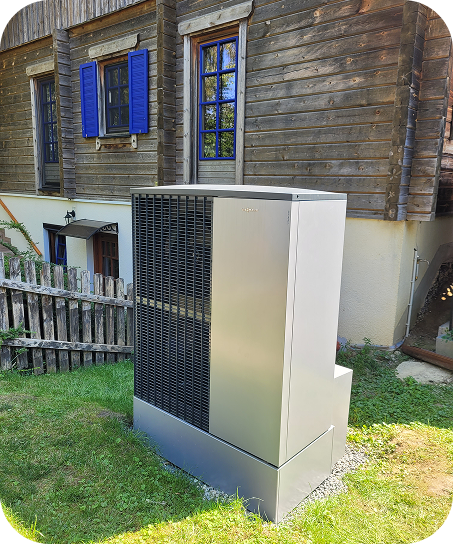 Moderne Wärmepumpe steht auf grünem Rasen vor einem Holzhaus mit blauen Fensterläden.