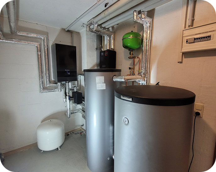 Technikraum mit Heizungsanlage, isolierten Rohrleitungen, zwei silbernen speicherbehältern und einem grünen Ausdehnungsgefäß an der Wand.